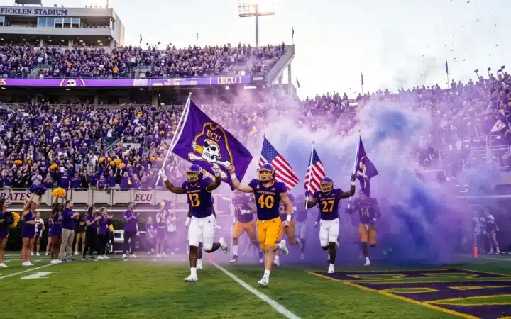 East Carolina University sports programs: Pirates football players run through purple smoke onto the field at Dowdy-Ficklen Stadium